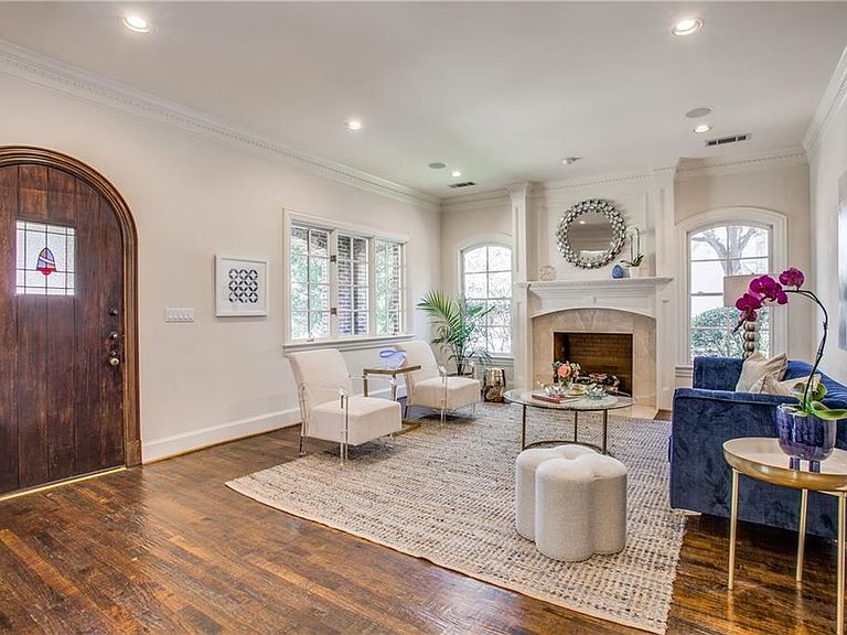 Entry living room of 4501 Livingston, with the arched wood front door inset with stained glass on the left, white walls and crown molding, a stone fireplace with mirrored over-mantel, and wide-plank hardwood floors