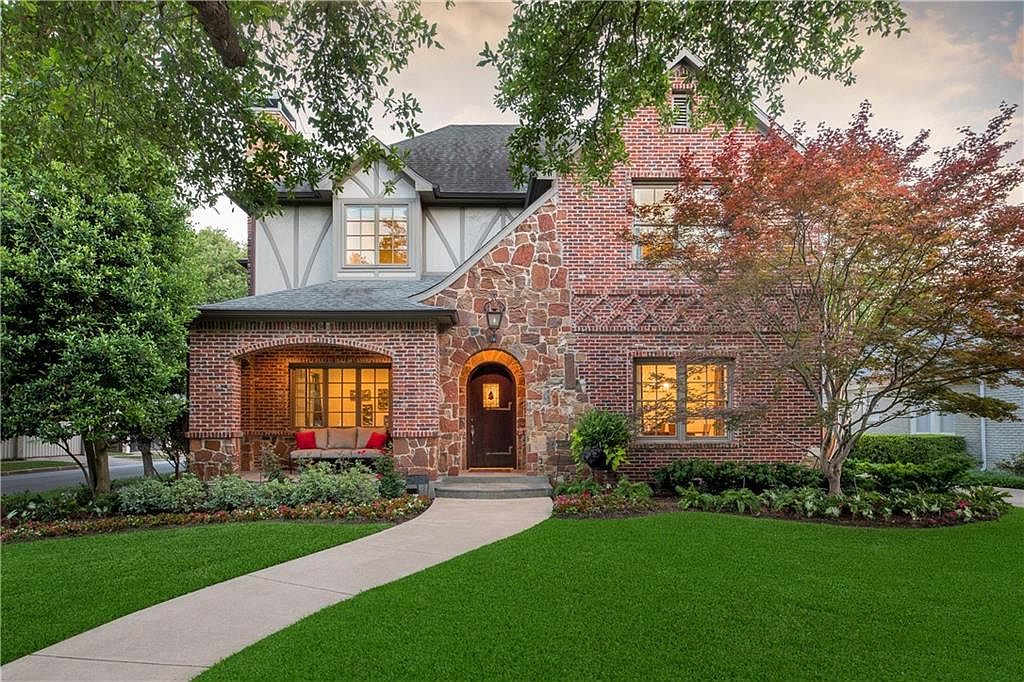 Front elevation of 4501 Livingston Ave, a two-story Tudor-influenced Highland Park residence in brick and rough-cut limestone with a wood arched door, leaded windows, and a covered porch lit at twilight